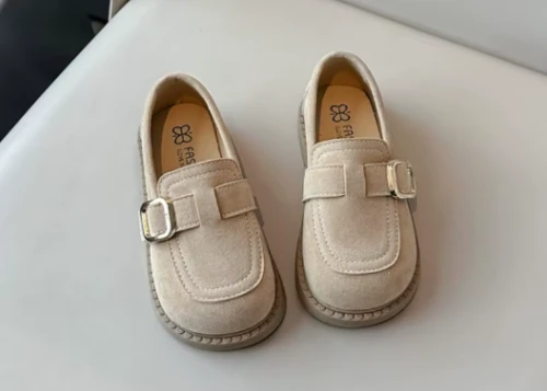 Zamszowe mokasyny dla dziewczynki z ozdobnym paskiem i prostym złotym elementem, dostępne w kolorze brązowym i beżowym.