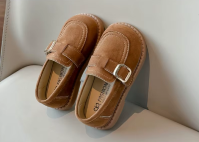 Zamszowe mokasyny dla dziewczynki z ozdobnym paskiem i prostym złotym elementem, dostępne w kolorze brązowym i beżowym.