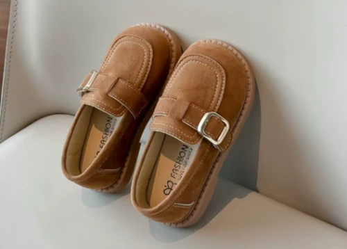 Zamszowe mokasyny dla dziewczynki z ozdobnym paskiem i prostym złotym elementem, dostępne w kolorze brązowym i beżowym.