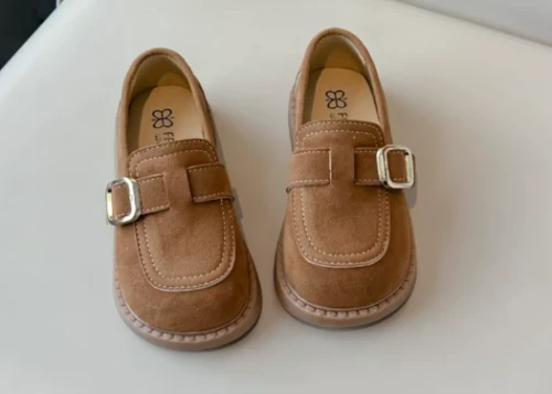 Zamszowe mokasyny dla dziewczynki z ozdobnym paskiem i prostym złotym elementem, dostępne w kolorze brązowym i beżowym.