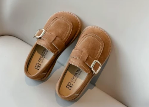 Zamszowe mokasyny dla dziewczynki z ozdobnym paskiem i prostym złotym elementem, dostępne w kolorze brązowym i beżowym.